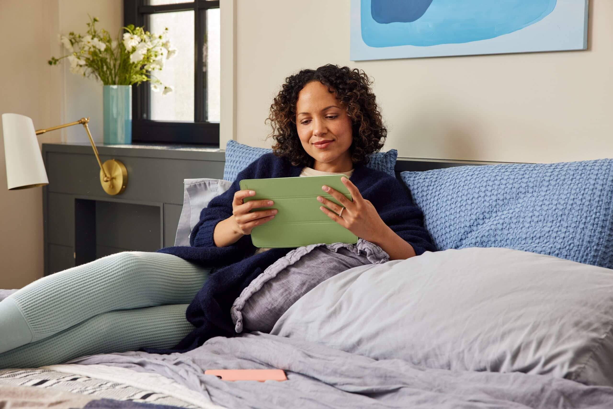 A woman with curly hair laying in bed looking at a tablet with a green screen protector