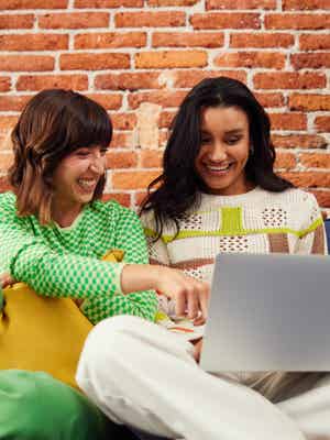 Dos amigas sonríen mirando una computadora, lo que ilustra un ejemplo de alguien que compra con PayPal.