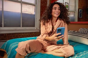 A woman sits on a bed, smiling as she holds a blue phone in her hands, enjoying a moment of connection.